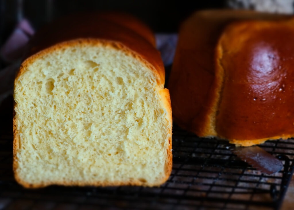 Ein weiches und fluffiges Brot auf einem schwarzen Gitter. Der Hintergrund ist dunkel, neben dem Brioche liegt eine abgeschnittene Hälfte des Brotes auf dem Brotmesser